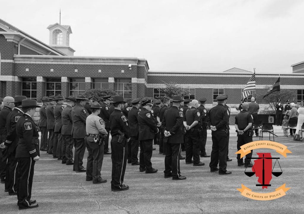 Police officers standing in rows at an event with an American flag an large building in the background. Logo on image: Kendall County Association of Chiefs of Police.